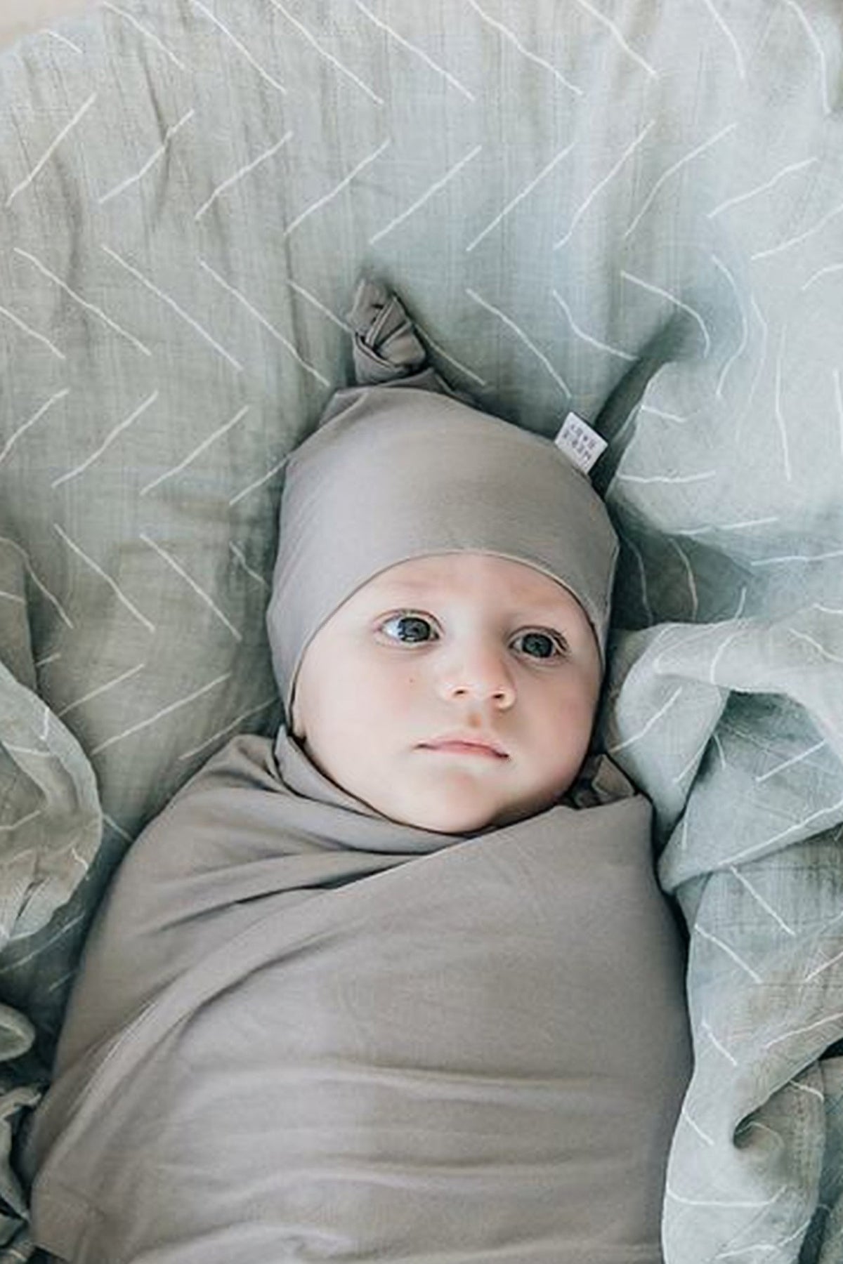 Newborn in a soft, gray bamboo knot hat and swaddle, lying on a light patterned blanket.