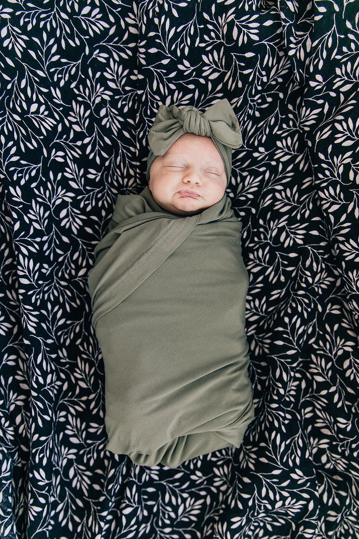 A baby wrapped in an olive green bamboo swaddle with a matching head wrap, lying on a leafy-patterned blanket.