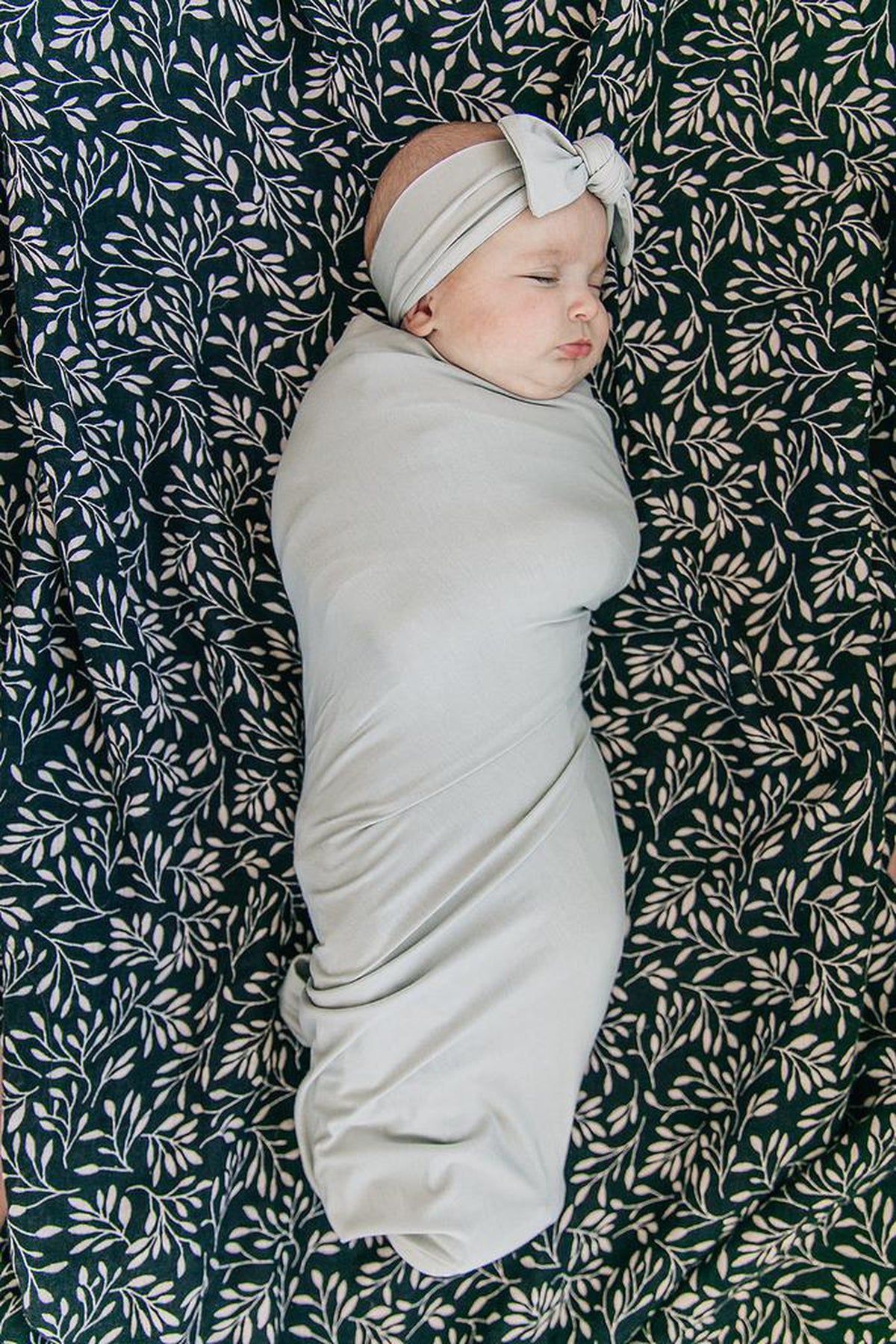 Sleeping baby wrapped in a soft, light gray swaddle with matching headband, lying on a leafy patterned blanket.