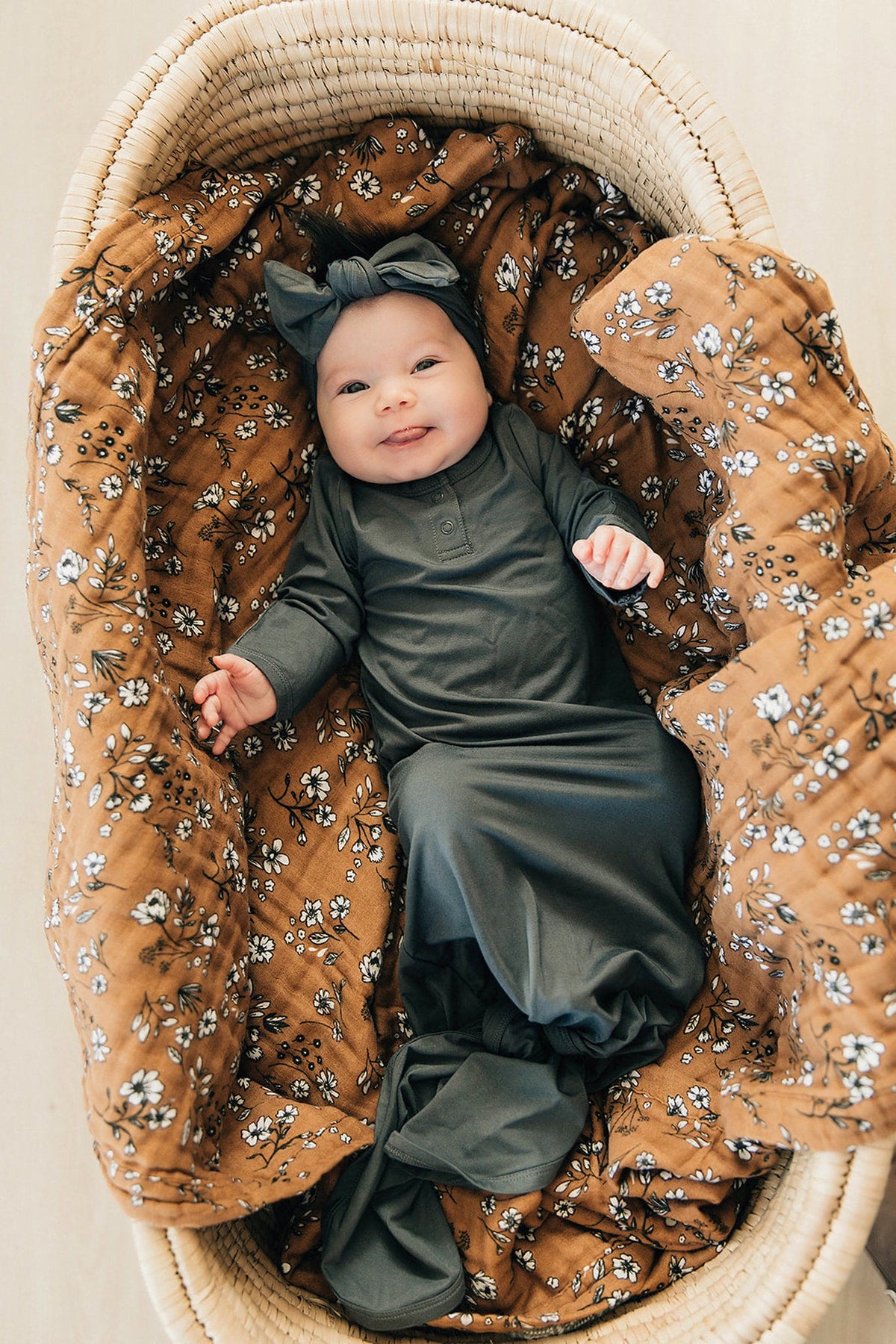 A baby in a charcoal knot gown lies in a wicker basket with a floral-patterned blanket.