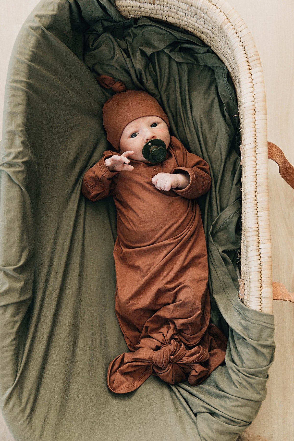 A baby in a rust-colored bamboo knot gown and matching hat lies in a woven bassinet with green bedding.