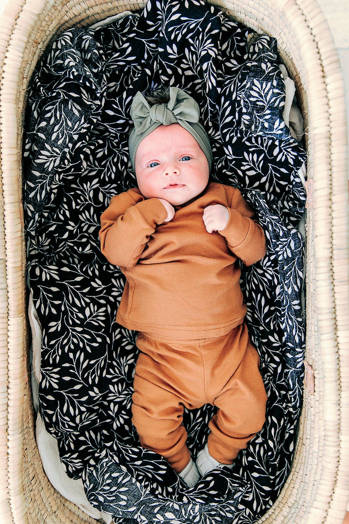 Baby in a brown outfit and olive head wrap lies in a basket with dark floral-patterned lining.