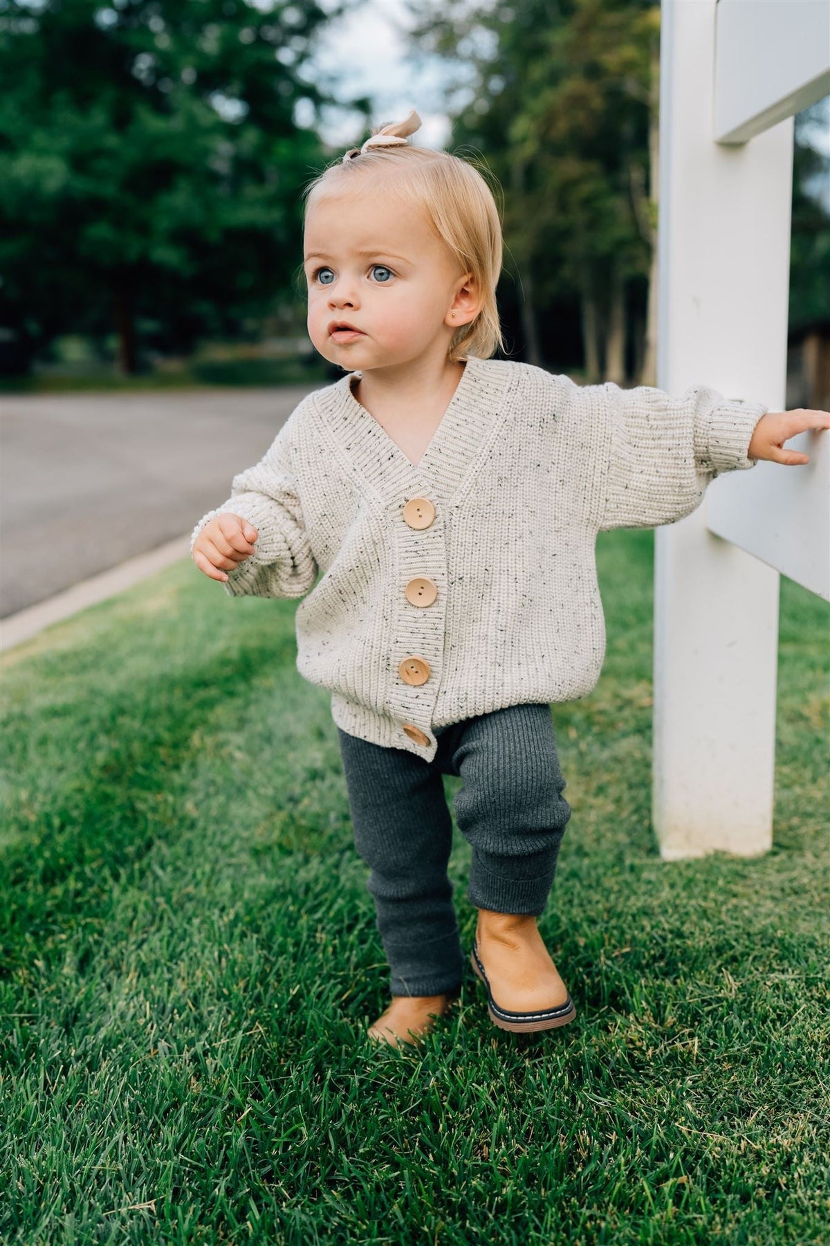 A toddler in a cozy knit sweater and charcoal leggings stands on grass, leaning against a white fence.