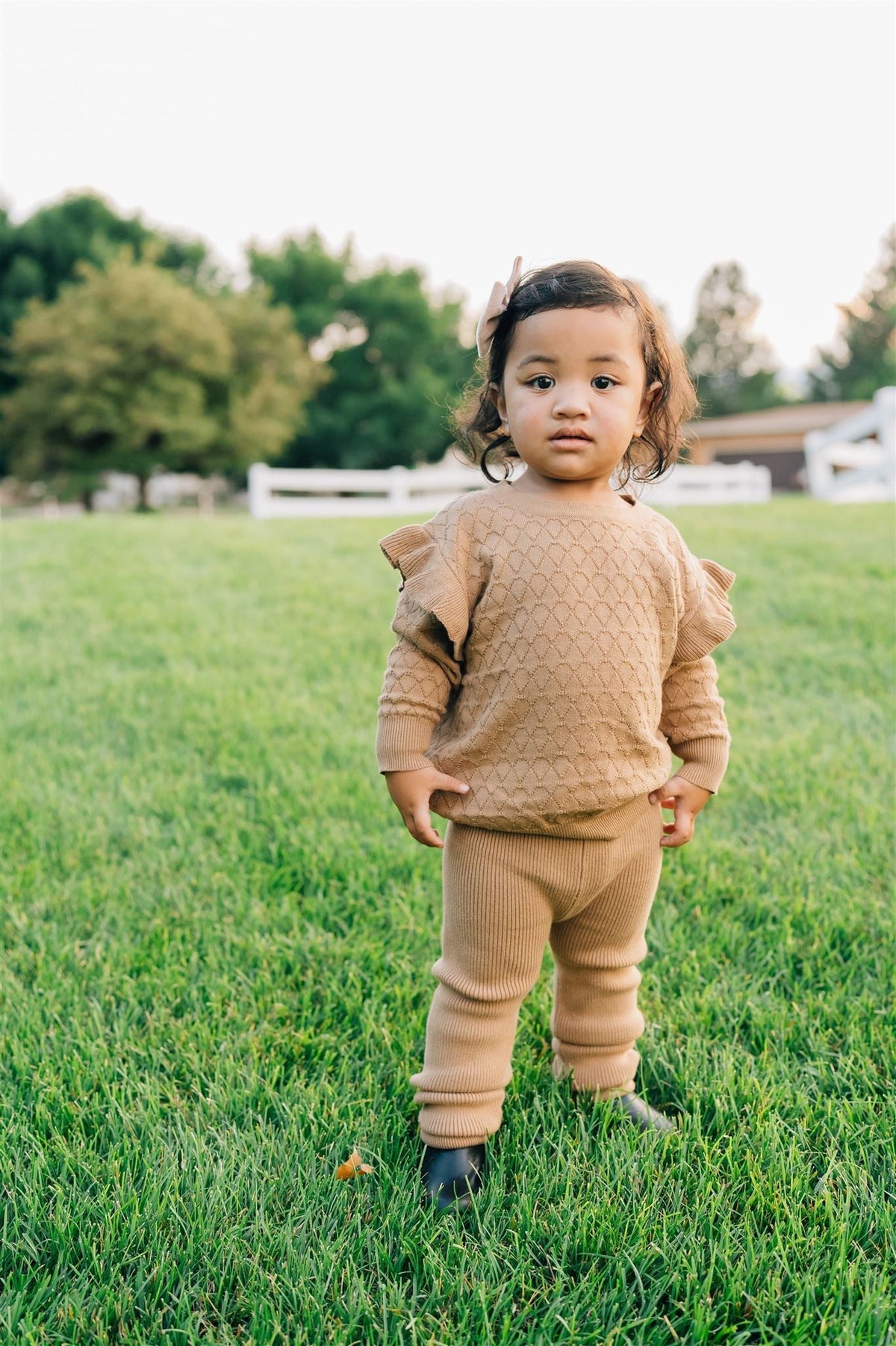 A toddler wearing toffee-colored knit leggings and a matching sweater stands on grass with trees in the background.