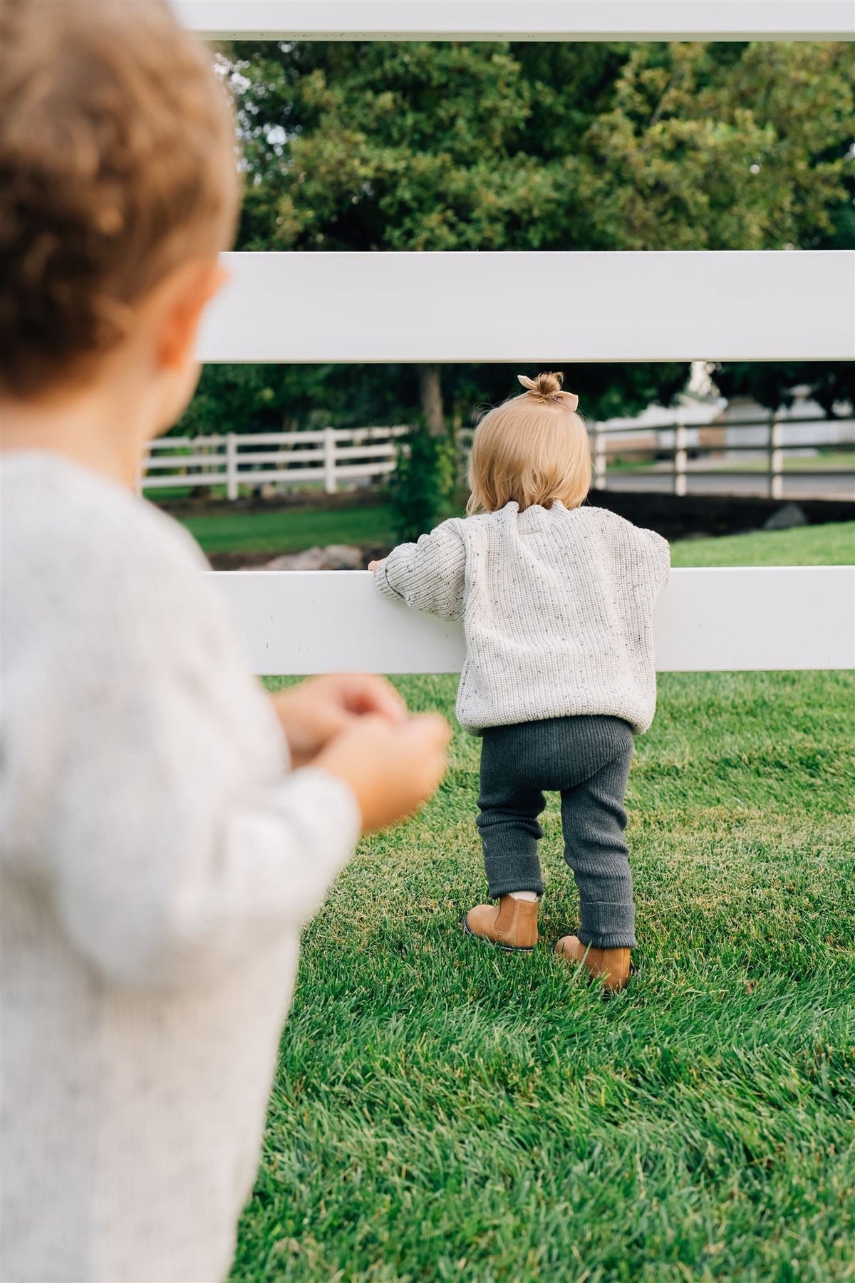 Two toddlers in a grassy area, one wearing charcoal knit leggings and a sweater, exploring near a white fence.