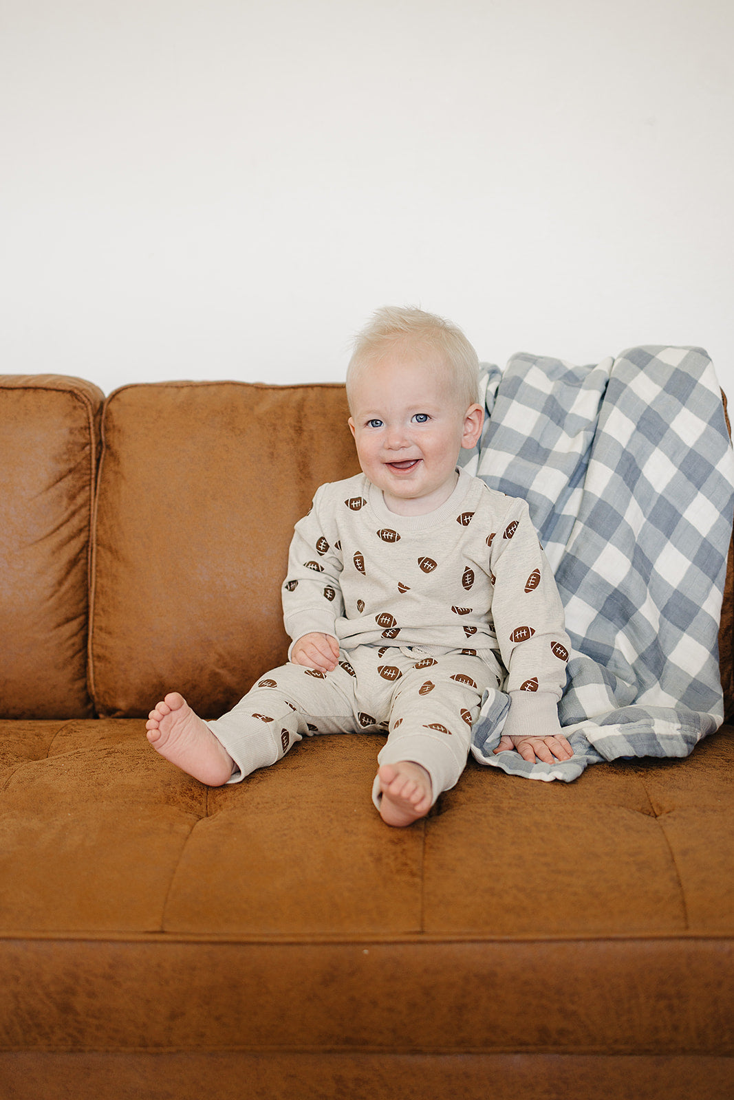 A smiling baby sits on a brown couch, wearing a light beige French terry cotton outfit with a playful football print. A blue and white checkered blanket is draped nearby.