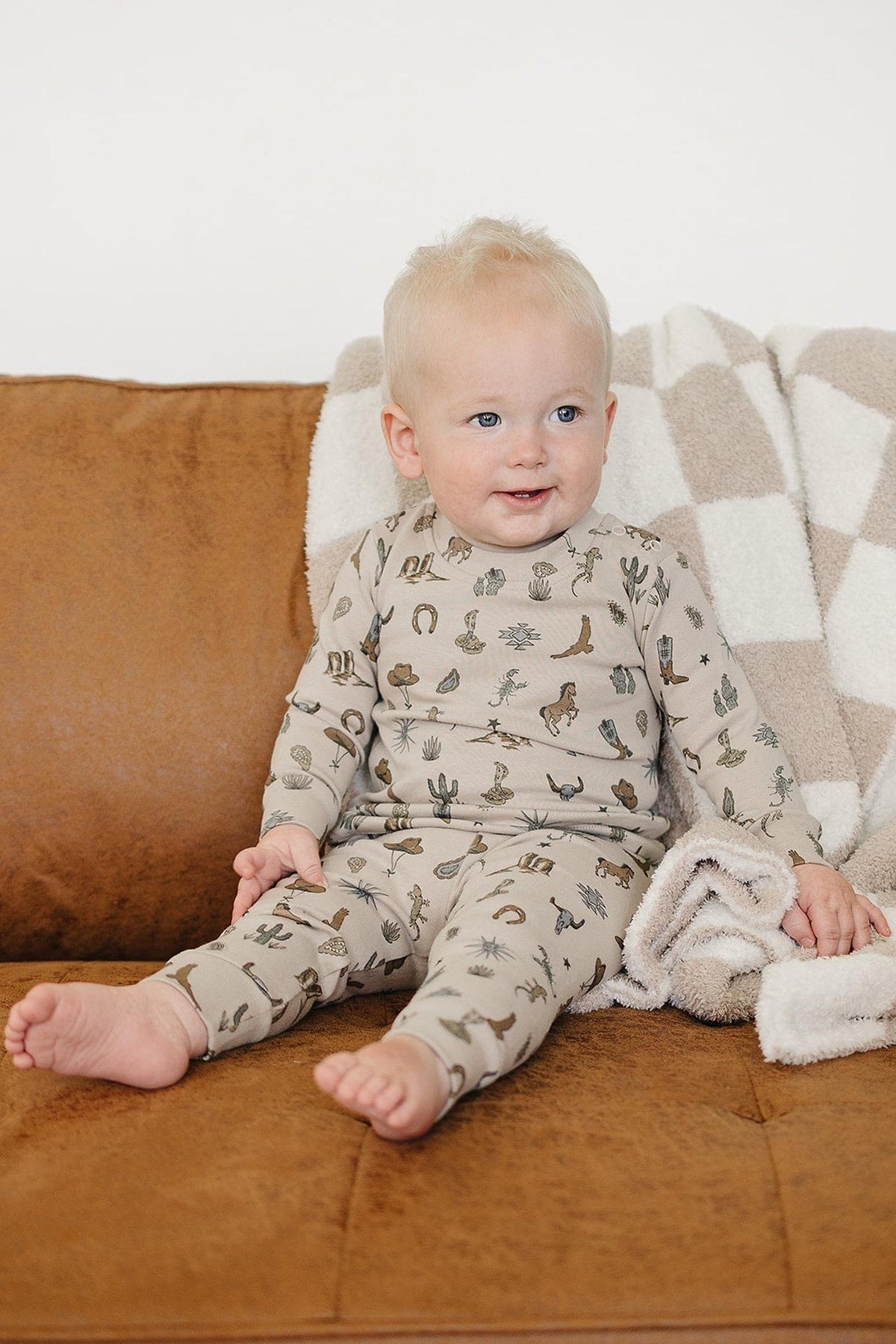 A smiling toddler sits on a brown couch, wearing a beige cotton outfit adorned with cactus patterns. A cozy, checkered blanket drapes over the couch, enhancing the warm atmosphere.