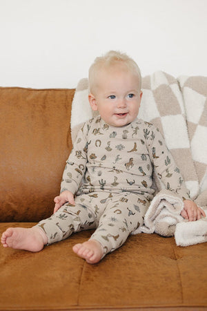 A smiling toddler sits on a brown couch, wearing a beige cotton outfit adorned with cactus patterns. A cozy, checkered blanket drapes over the couch, enhancing the warm atmosphere.