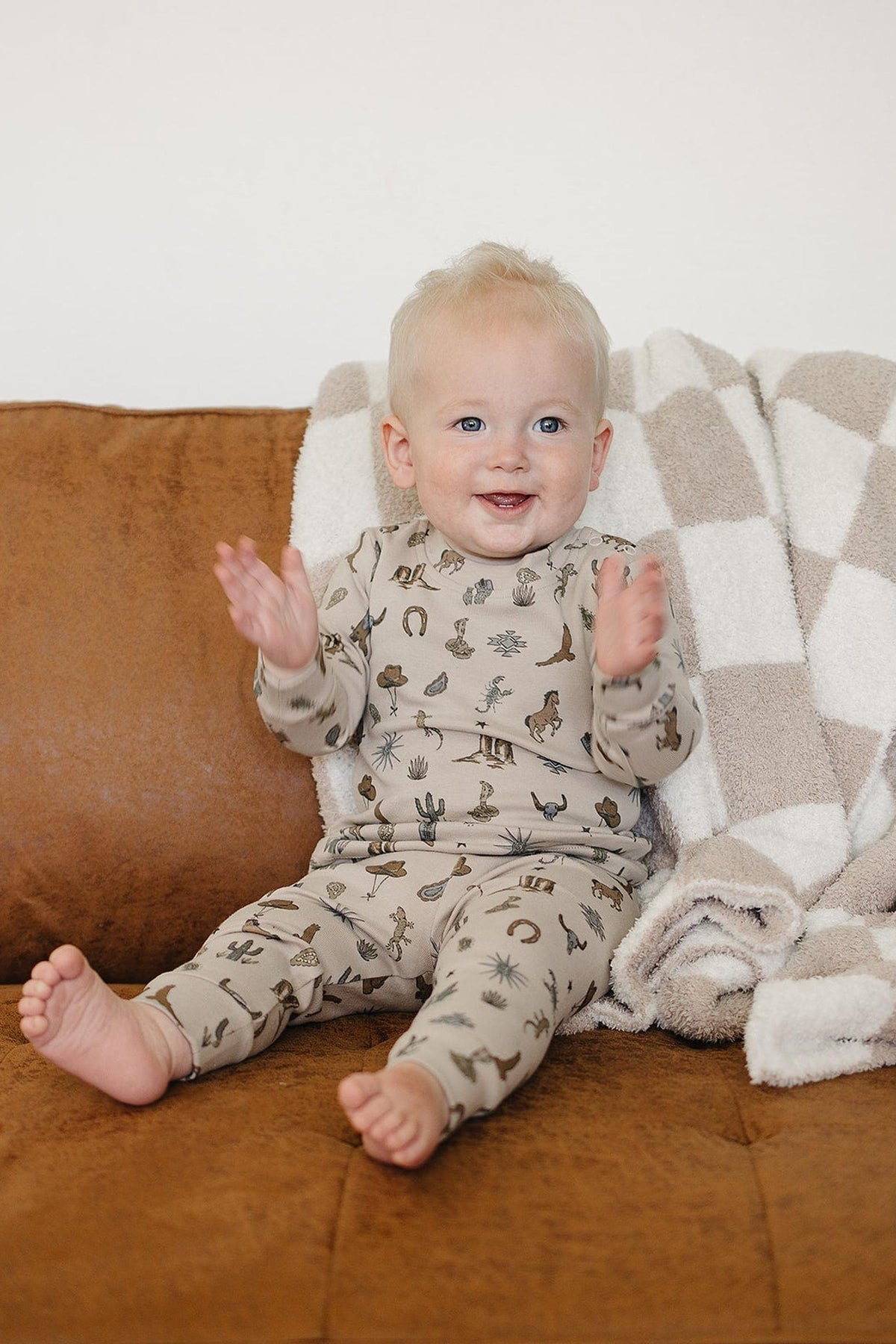 A smiling toddler sits on a brown couch, wearing a beige cotton outfit with whimsical animal prints. A soft, checkered blanket is draped over the couch, enhancing the cozy atmosphere.