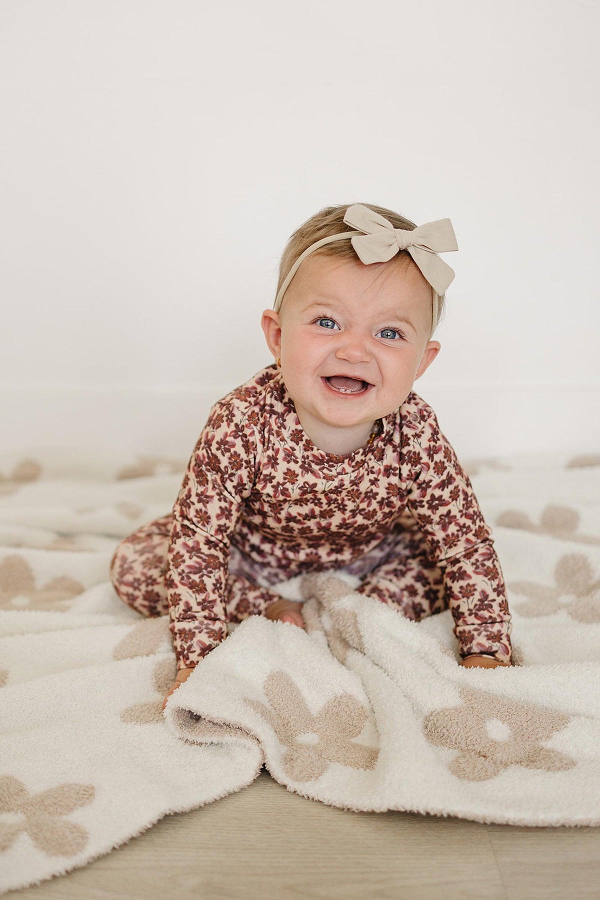 A joyful baby sits on a soft, light-colored blanket, wearing an Antique Floral Cotton Set featuring a long sleeve top with a floral pattern.