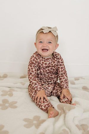A joyful baby sits on a soft, patterned blanket, wearing a long-sleeve, antique floral cotton outfit featuring earthy tones. The child has a light bow headband, enhancing the adorable look.