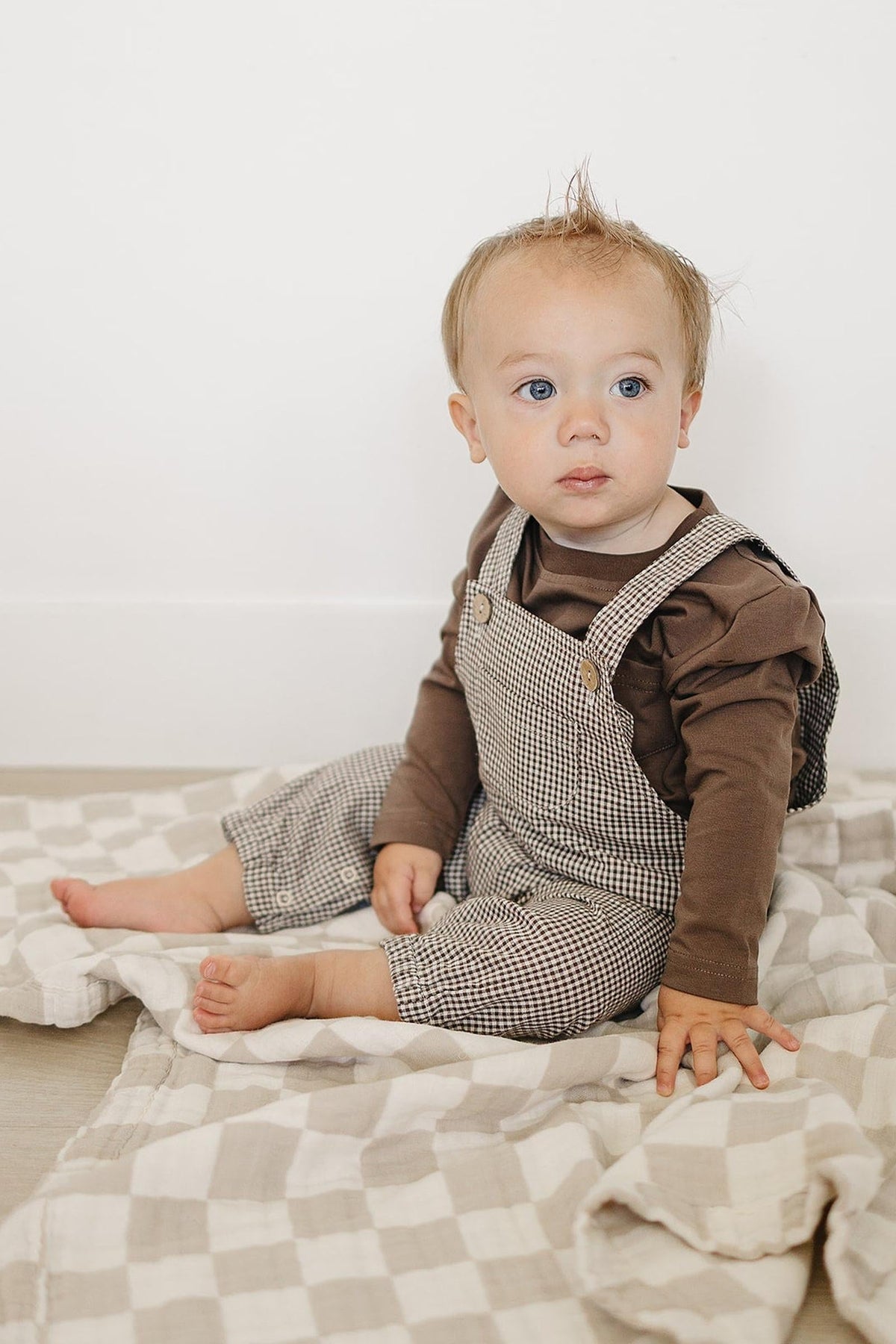 A toddler sits on a checkered blanket, wearing a brown long sleeve shirt and checkered overalls. The child gazes thoughtfully, showcasing a relaxed and playful outfit suitable for everyday wear.