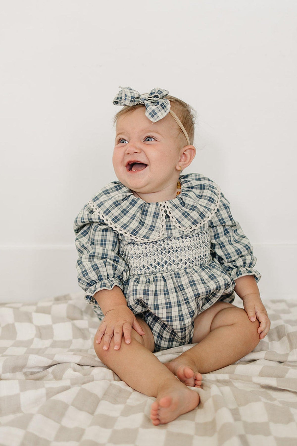 A joyful baby sits on a checkered blanket, wearing a blue plaid bubble romper with a ruffle collar. The romper features long sleeves and is made from soft, breathable cotton.