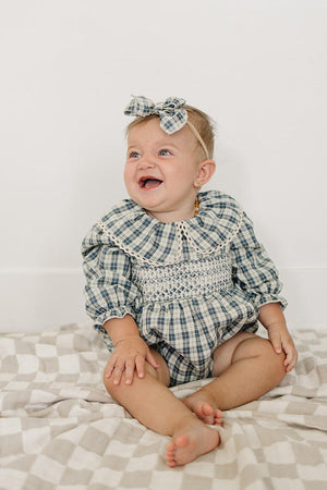 A joyful baby sits on a checkered blanket, wearing a blue plaid bubble romper with a ruffle collar. The romper features long sleeves and is made from soft, breathable cotton.