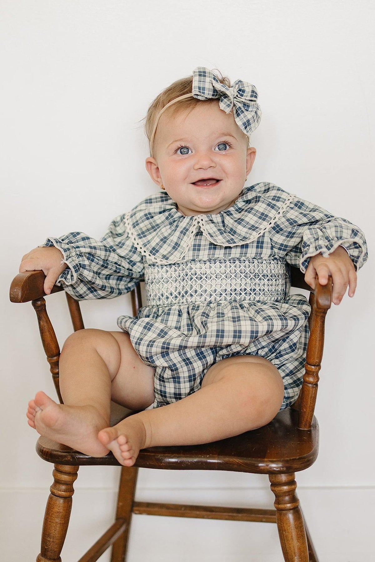 A smiling baby sits on a vintage wooden chair, wearing a blue plaid romper with a ruffle collar and matching hair bow. The romper features soft, breathable cotton for comfort.