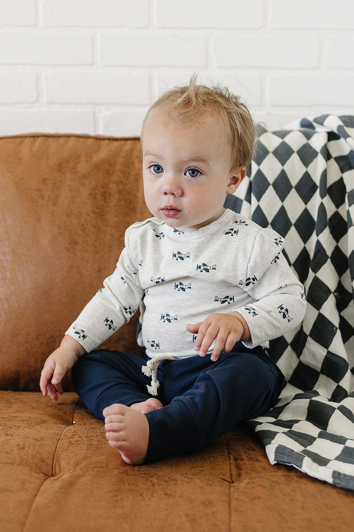 A young child sits on a brown couch, wearing a light gray long-sleeve shirt with airplane prints and navy pants. A black and white checkered blanket is draped nearby.