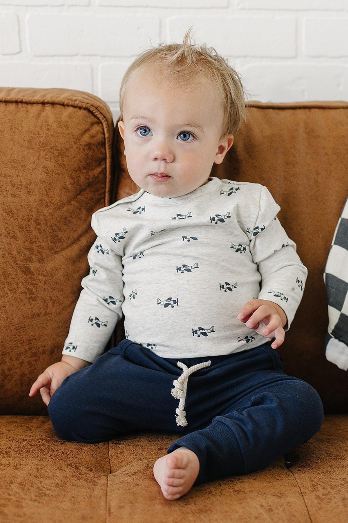 A toddler sits on a brown couch, wearing a light gray long-sleeve shirt with a playful print and navy cotton joggers. The cozy setting features a patterned throw blanket nearby.