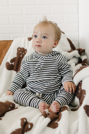 A baby sits on a brown leather couch, wearing a charcoal stripe ribbed hooded set. The soft outfit features a cozy hood and matching pants, complemented by a plush teddy.