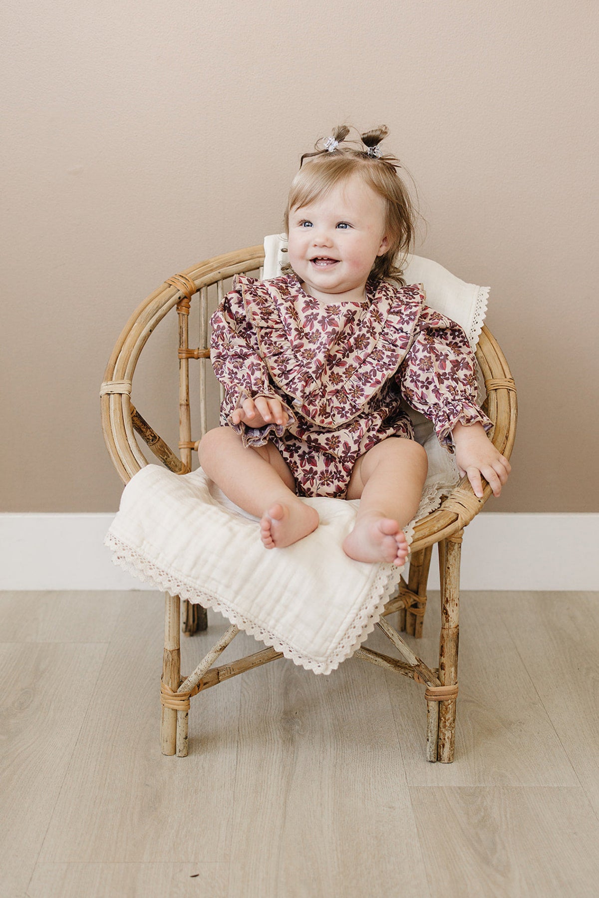 A joyful baby girl sits in a bamboo chair, wearing a vintage floral long sleeve cotton romper with ruffled details. The soft, breathable fabric and relaxed fit ensure comfort.