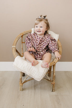 A joyful baby girl sits in a bamboo chair, wearing a vintage floral long sleeve cotton romper with ruffled details. The soft, breathable fabric and relaxed fit ensure comfort.