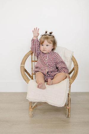 A baby girl with light brown hair is seated in a bamboo chair, wearing a red gingham cotton outfit with a Peter Pan collar. She raises one hand playfully.