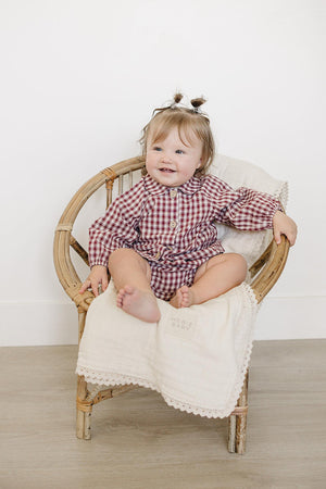 A smiling baby girl sits in a rattan chair, wearing a red gingham long-sleeve shirt and matching bloomer set.
