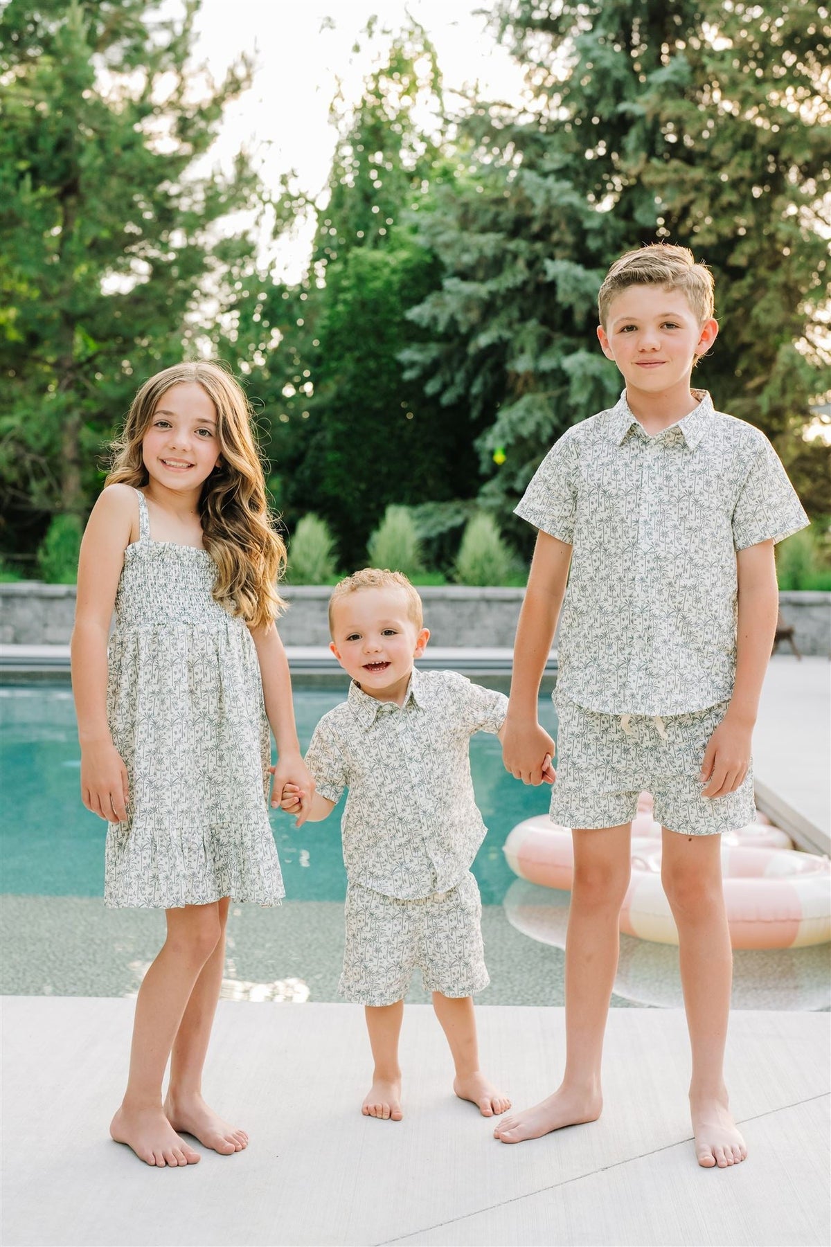 Three children in matching palm tree print outfits stand barefoot by a pool, surrounded by lush greenery.