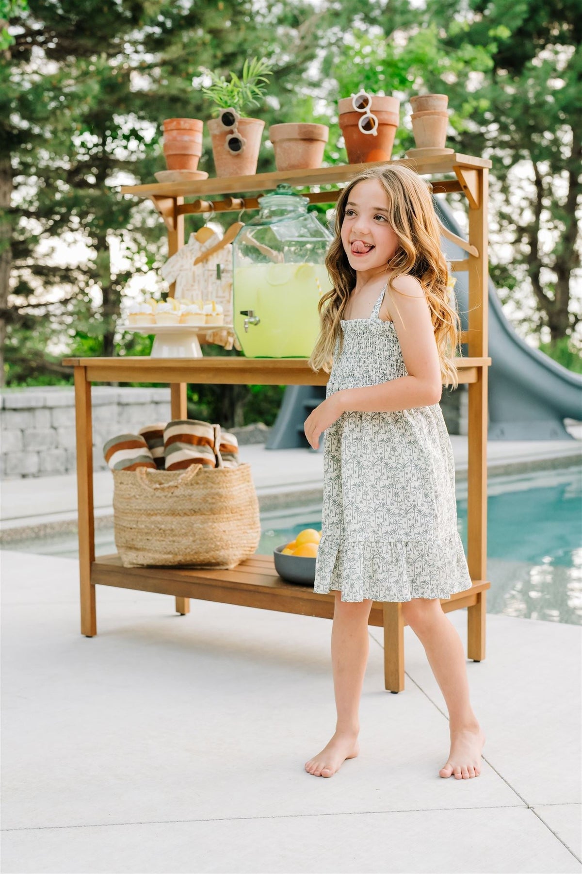 A young girl in a palm tree patterned dress stands barefoot by a poolside lemonade stand.