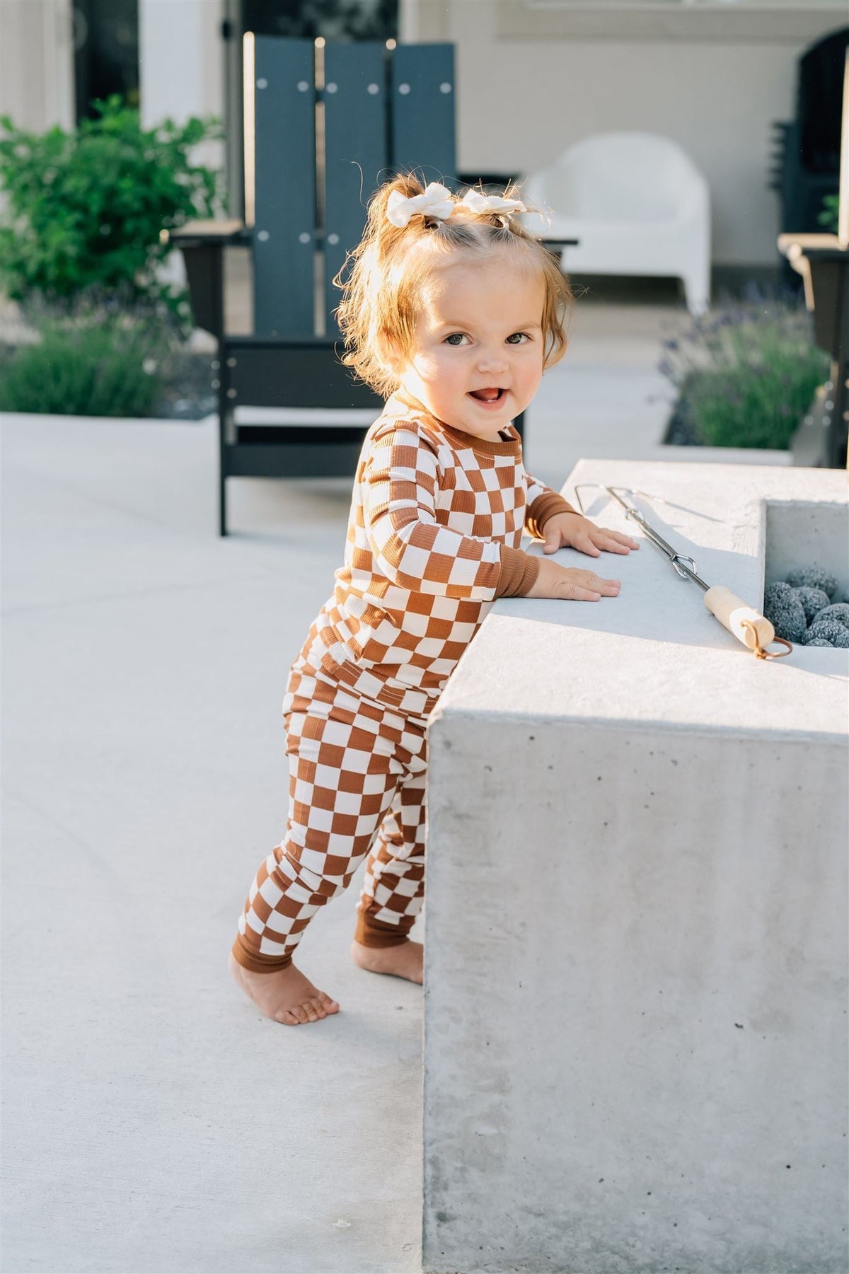 A toddler wearing a rust checkered pajama set stands outdoors, smiling near a concrete fire pit.