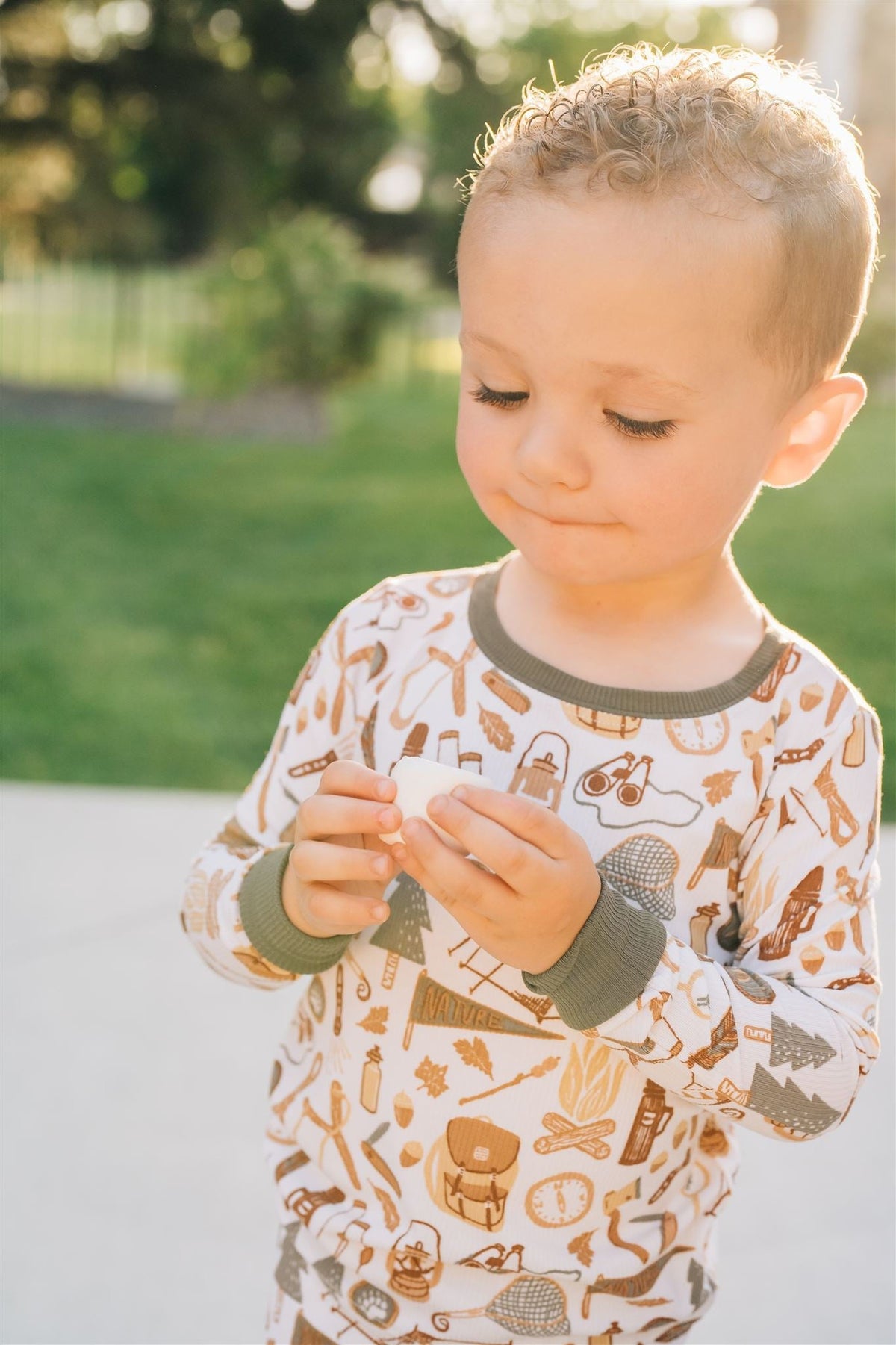 A toddler in camping-themed pajamas with green cuffs holds a small object outdoors on a sunny day.