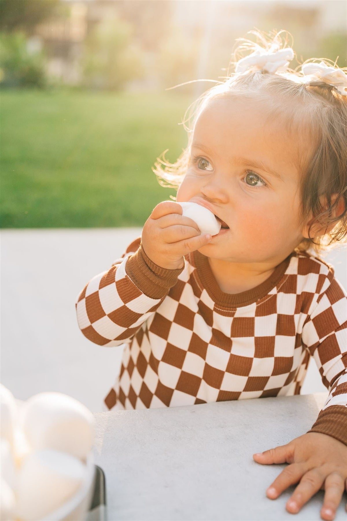A child in a rust checkered pajama set holds a white egg outdoors in soft sunlight.