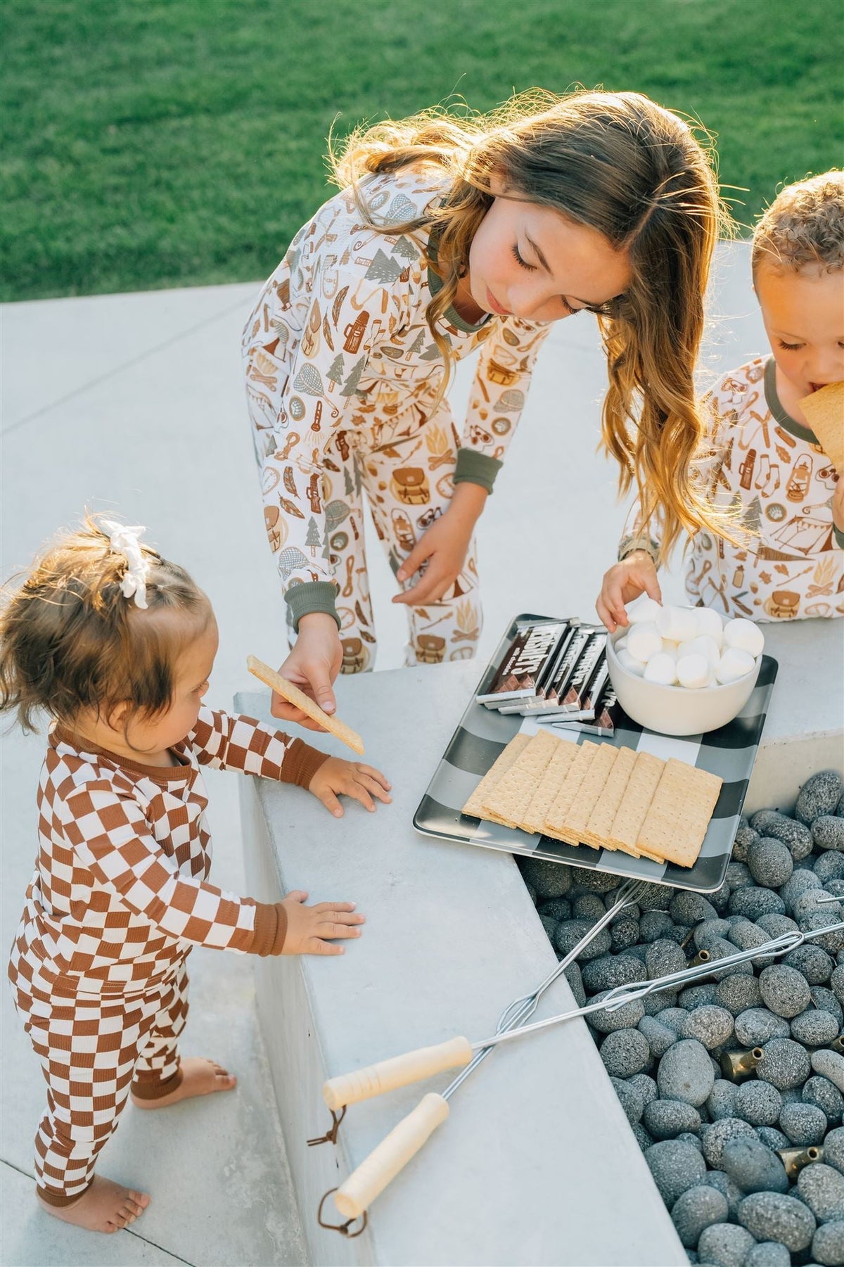 Children in matching pajamas prepare smores outdoors, with a tray of graham crackers, chocolate, and marshmallows nearby.