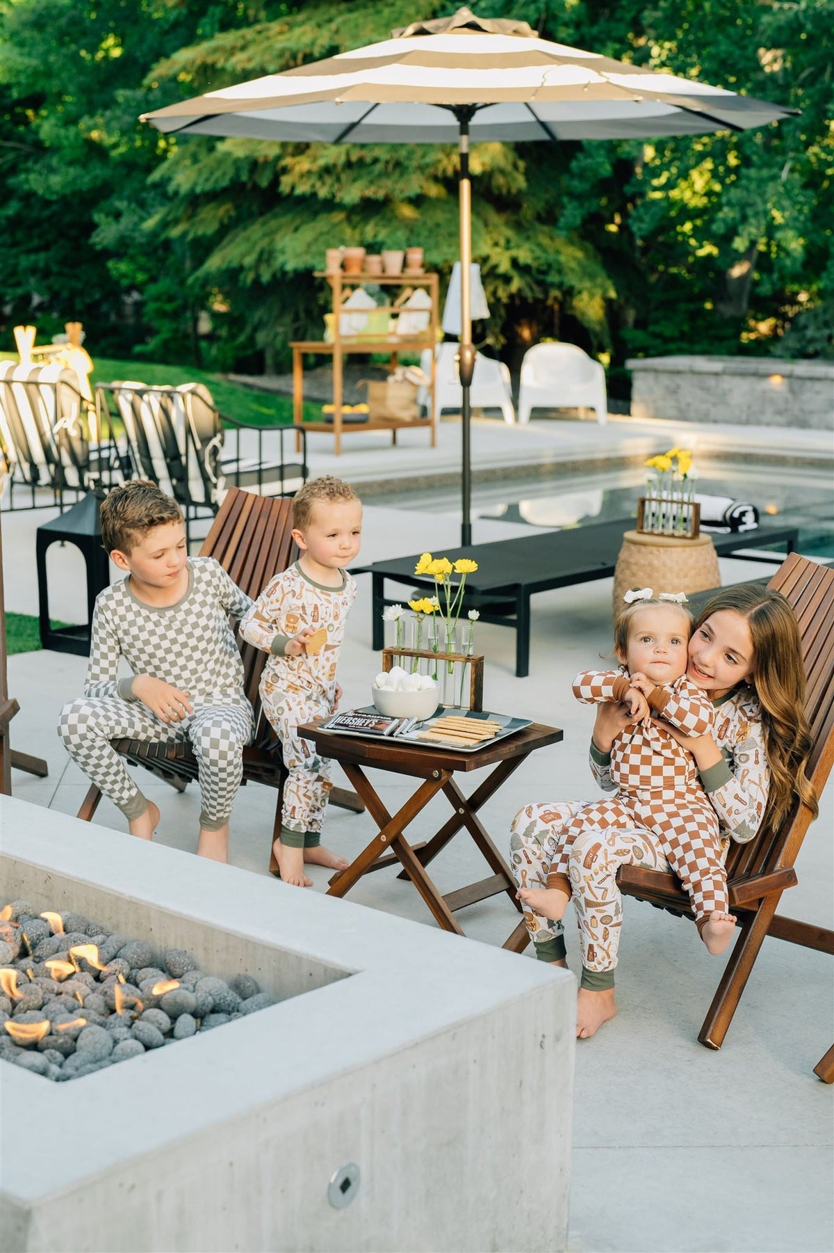 Children in checkered pajamas sit by a poolside fire pit, enjoying snacks and outdoor seating under an umbrella.