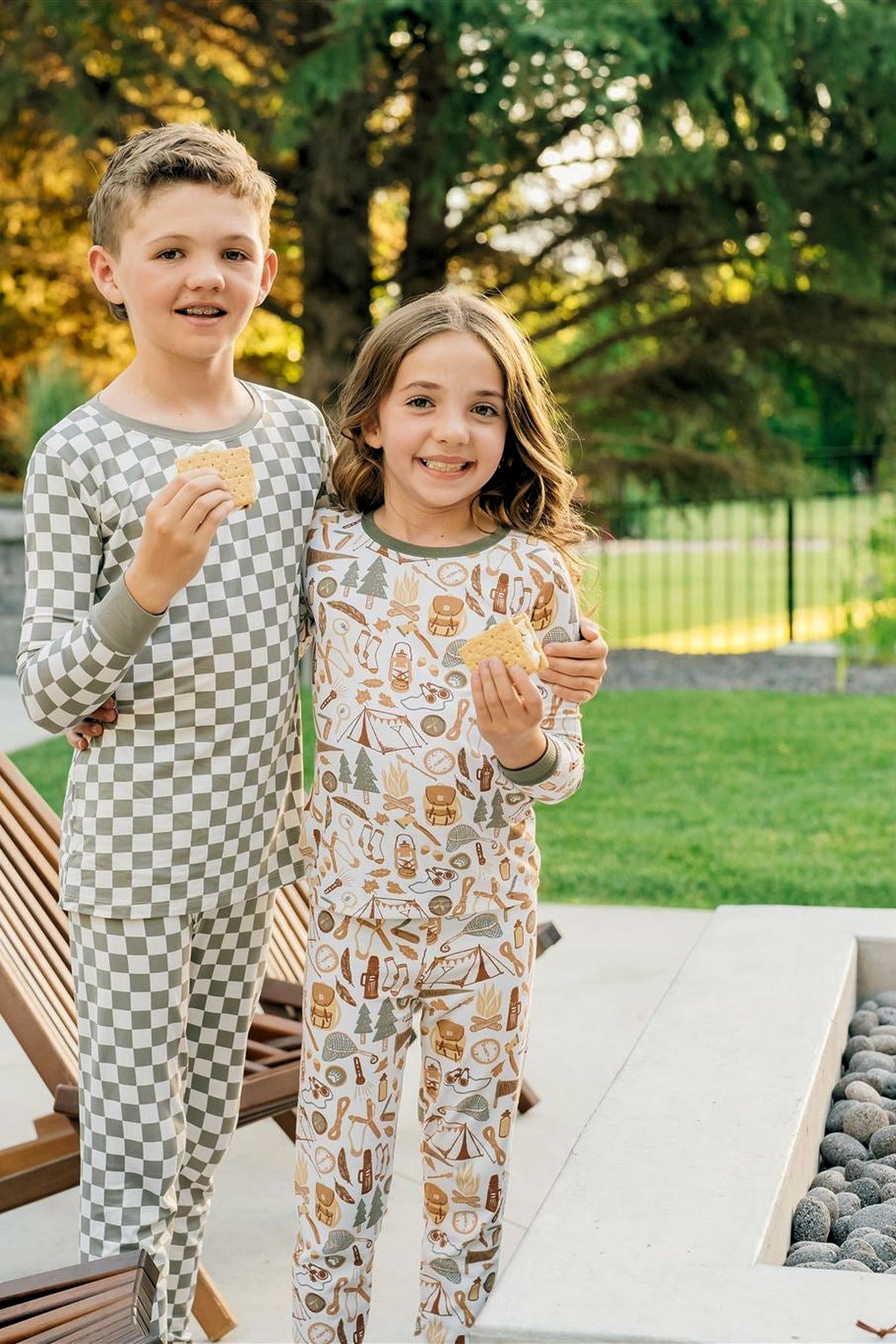 Two children outdoors wearing patterned pajamas; one in checkered, the other with camping motifs, holding snacks.