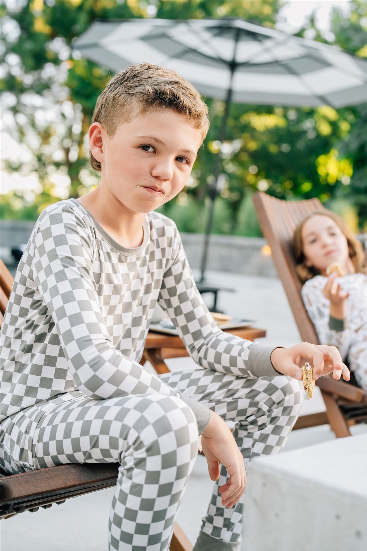 A child wearing a light green checkered pajama set sits outdoors, holding a snack in hand.