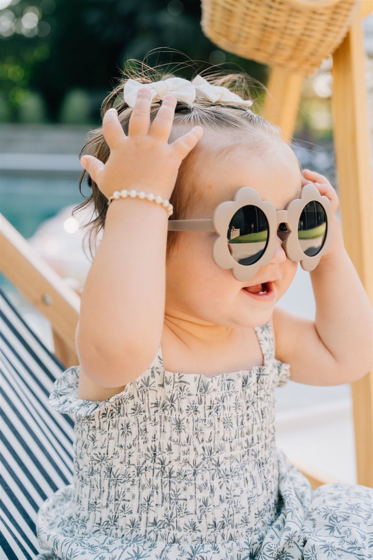 A toddler wearing flower-shaped sunglasses and a palm tree print dress smiles while sitting outdoors on a sunny day.