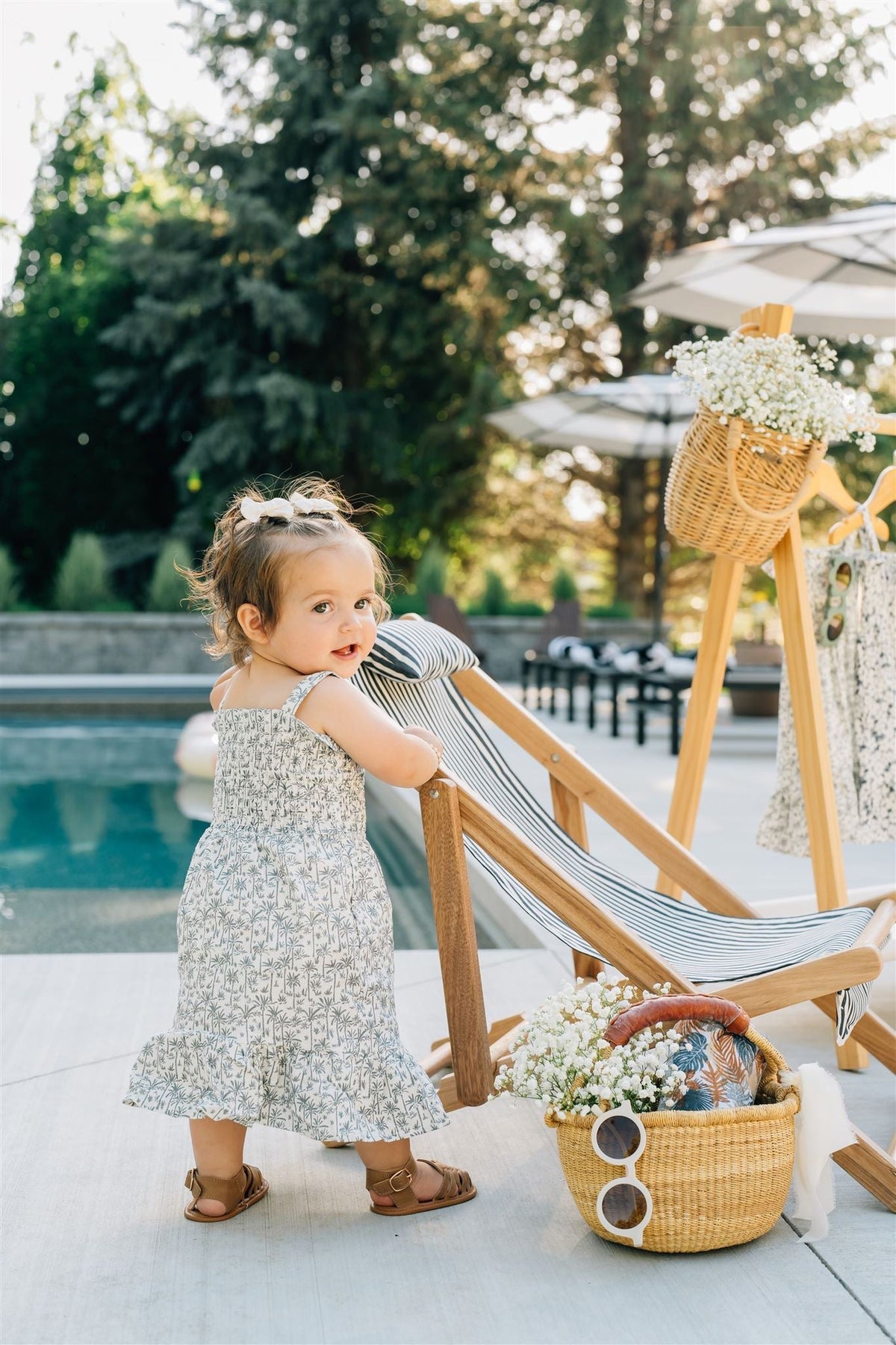 Toddler in a palm tree print dress stands by a wooden lounge chair near a pool, surrounded by greenery.