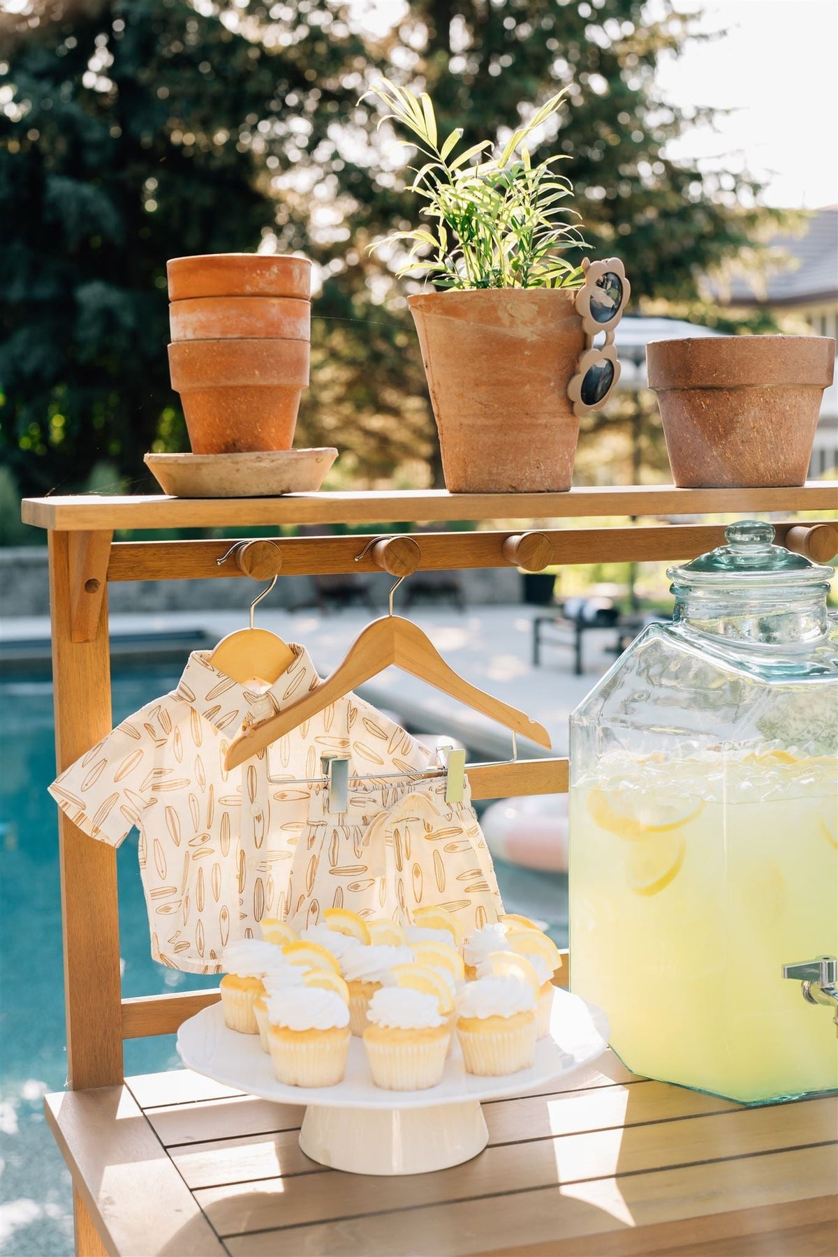 Wooden rack with surfboard-patterned baby shirt, shorts, terracotta pots, lemon cupcakes, and lemonade by a pool.