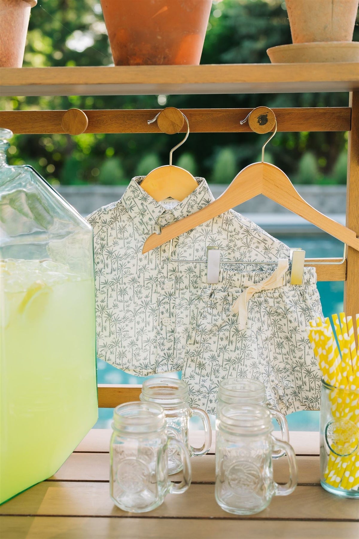 A palm tree patterned linen shirt and shorts set hangs on wooden hangers, with lemonade and mason jars nearby.