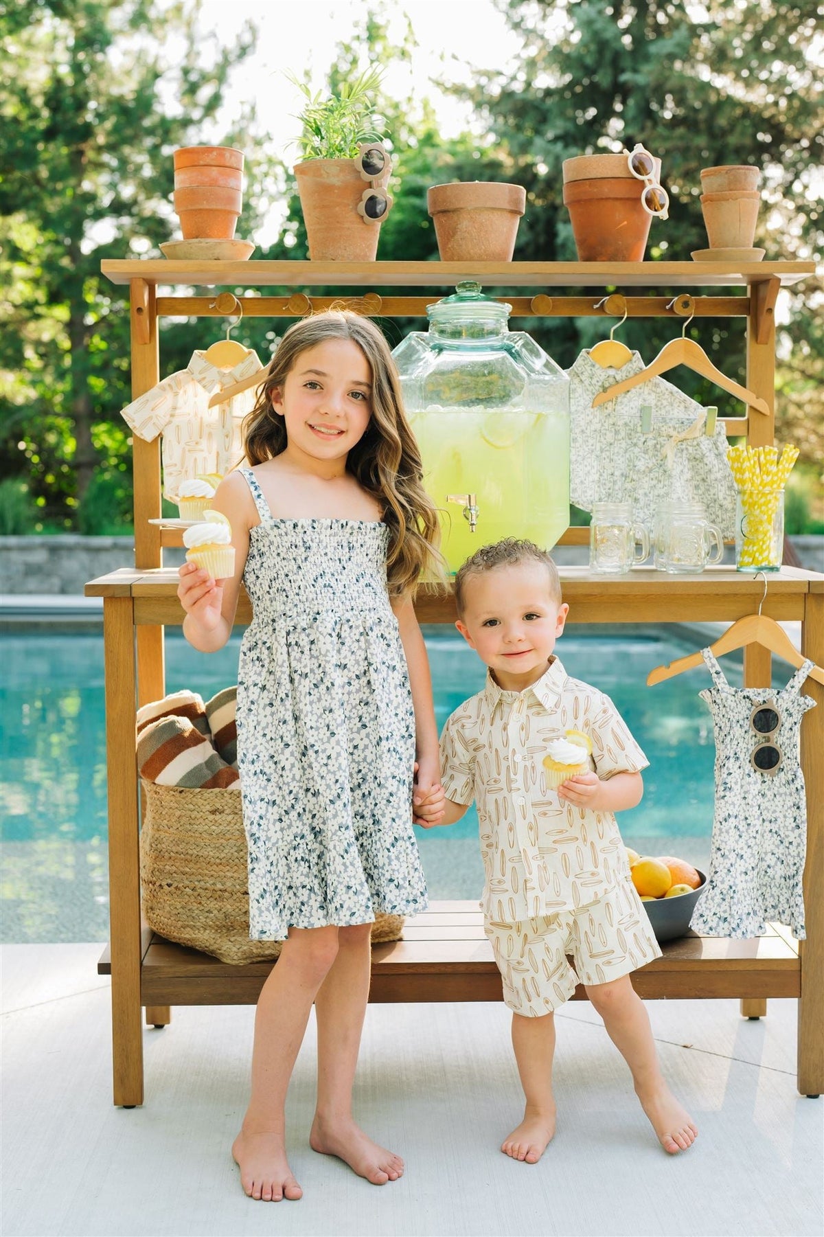 Children in summer outfits holding cupcakes by a poolside lemonade stand with hanging clothes and potted plants.