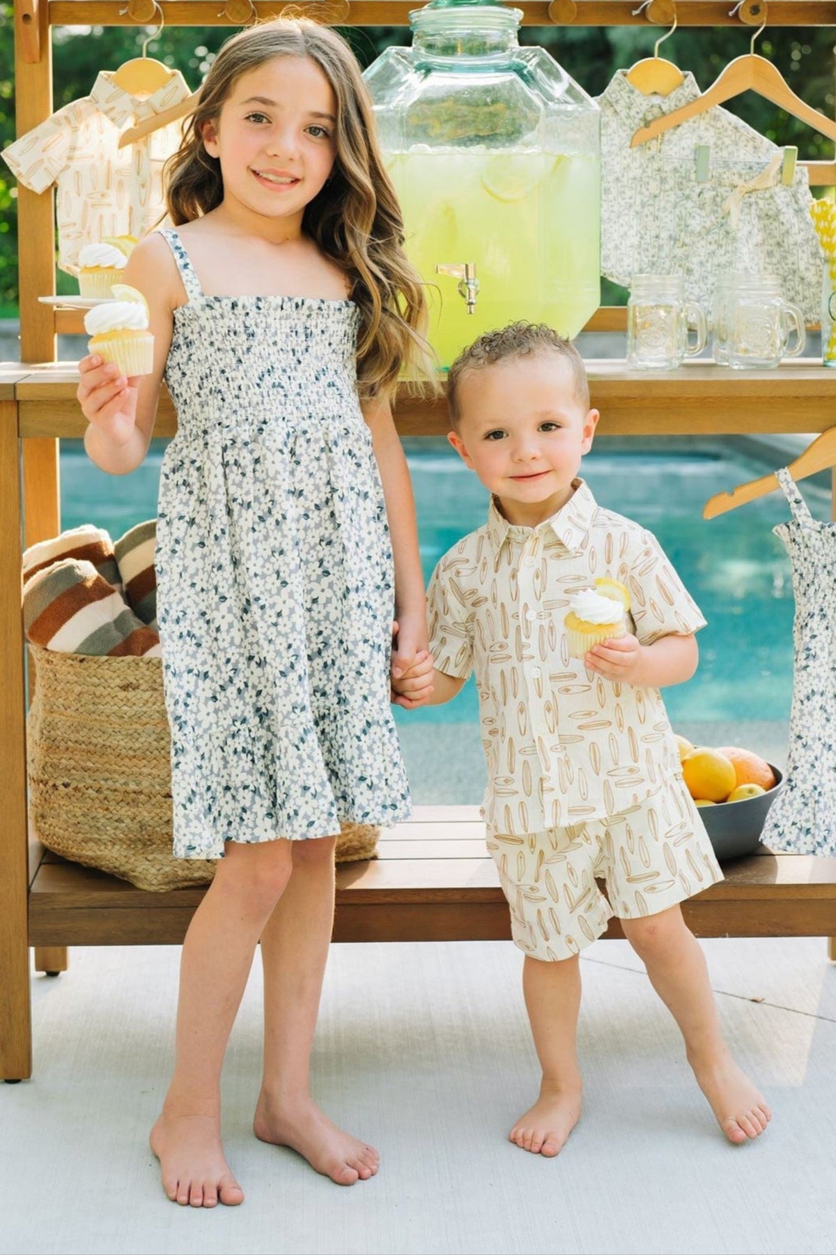 Two children stand by a lemonade stand, holding cupcakes, wearing summer outfits with surfboard and floral patterns.