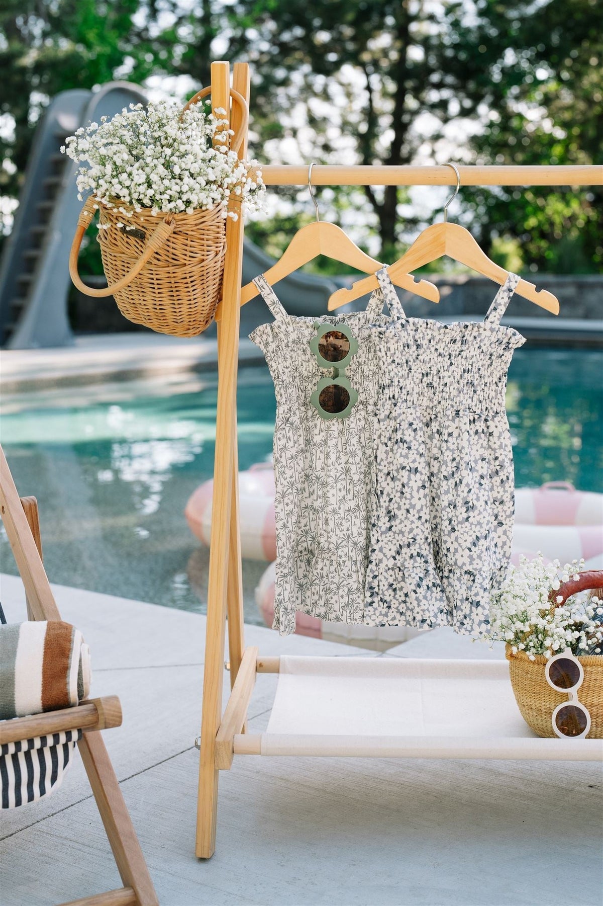 Two linen summer dresses with floral prints hang on wooden hangers by a poolside, next to a wicker basket.