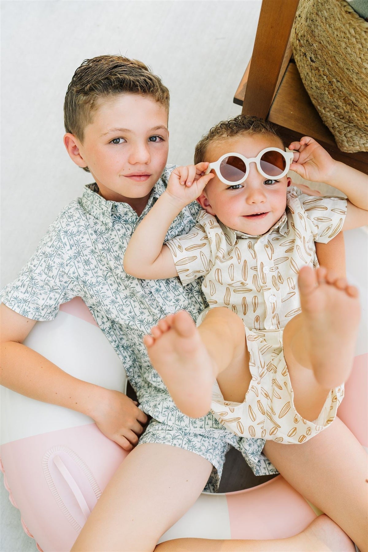 Two children wearing patterned button-up shirts, one with a surfboard print, sitting on an inflatable pool float.