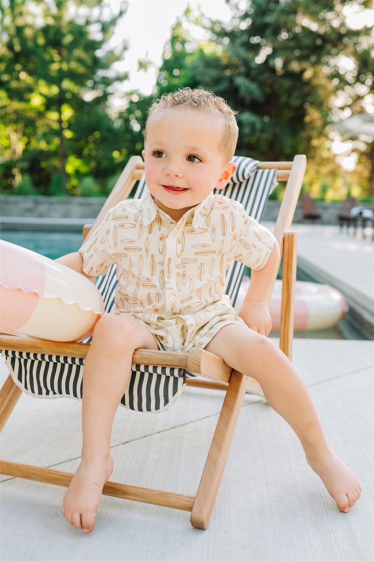 A toddler in a surfboard-patterned button-up shirt sits on a striped deck chair by the pool.