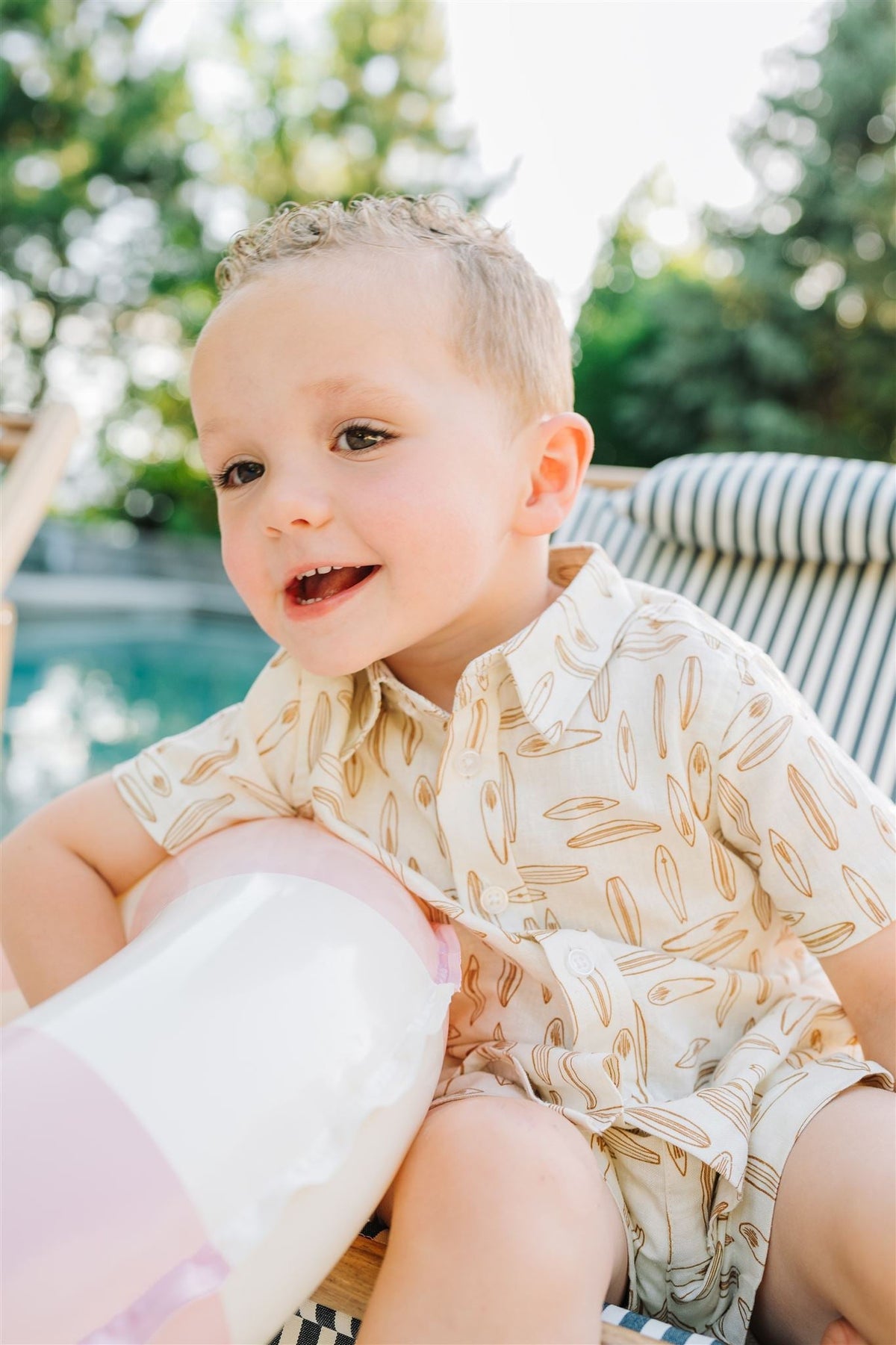 A young child wearing a beige patterned shirt smiles while sitting outdoors near a pool, holding an inflatable toy.