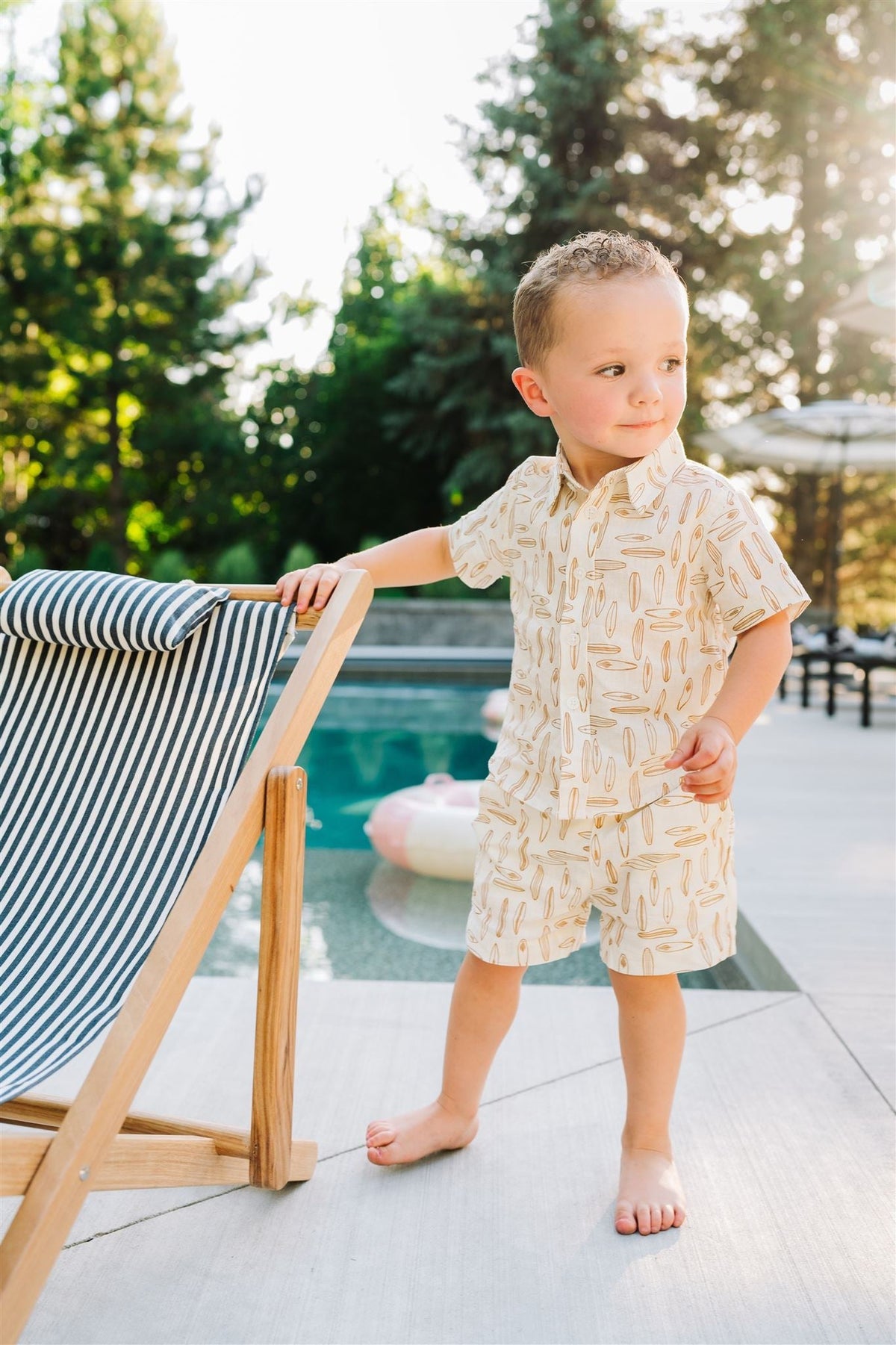 A child wearing a surfboard-patterned button-up shirt stands by a pool, holding a striped deck chair.