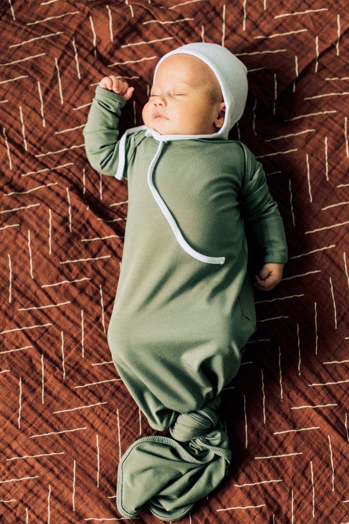 A baby sleeps on a rust-colored mudcloth muslin blanket, wearing a green outfit and white cap.