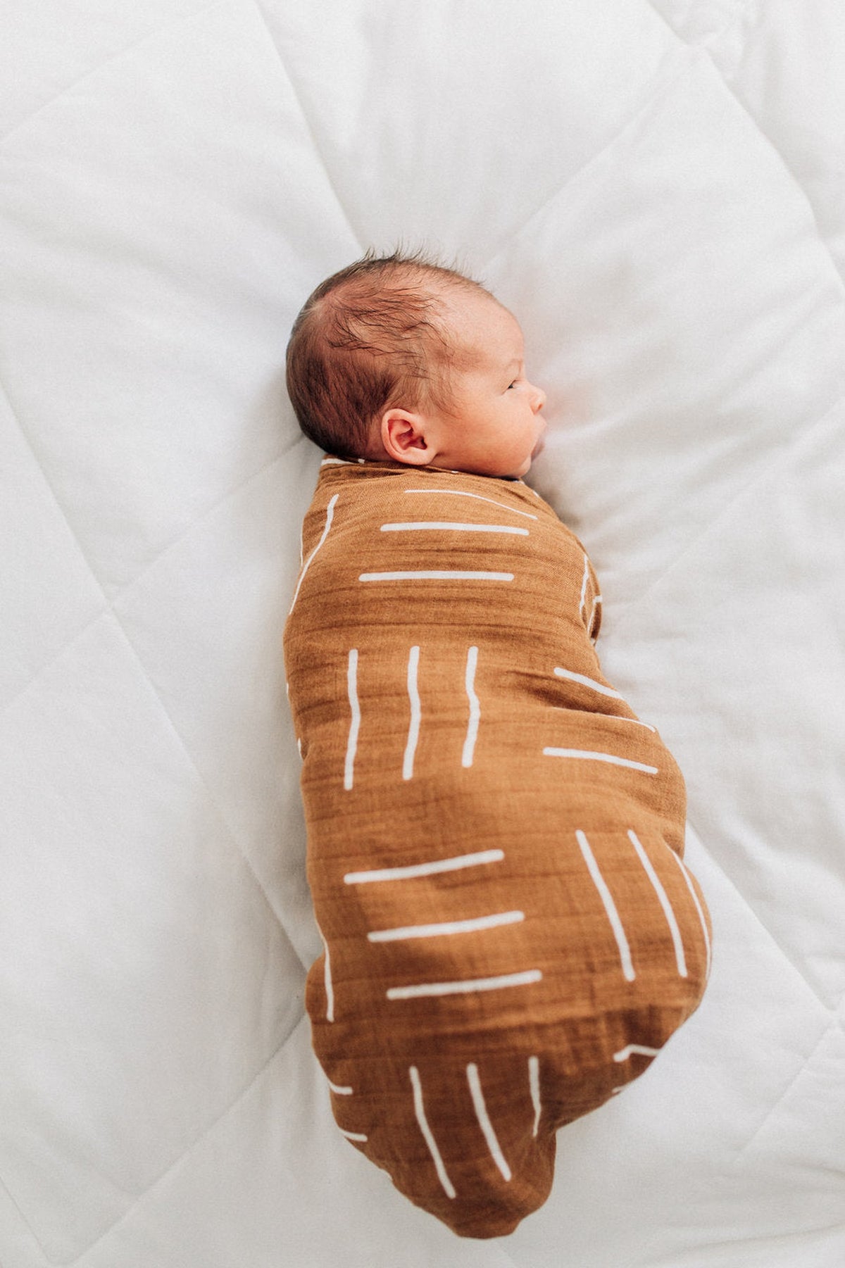 A baby is wrapped in a mustard-colored muslin swaddle with white geometric patterns, lying on a white quilt.
