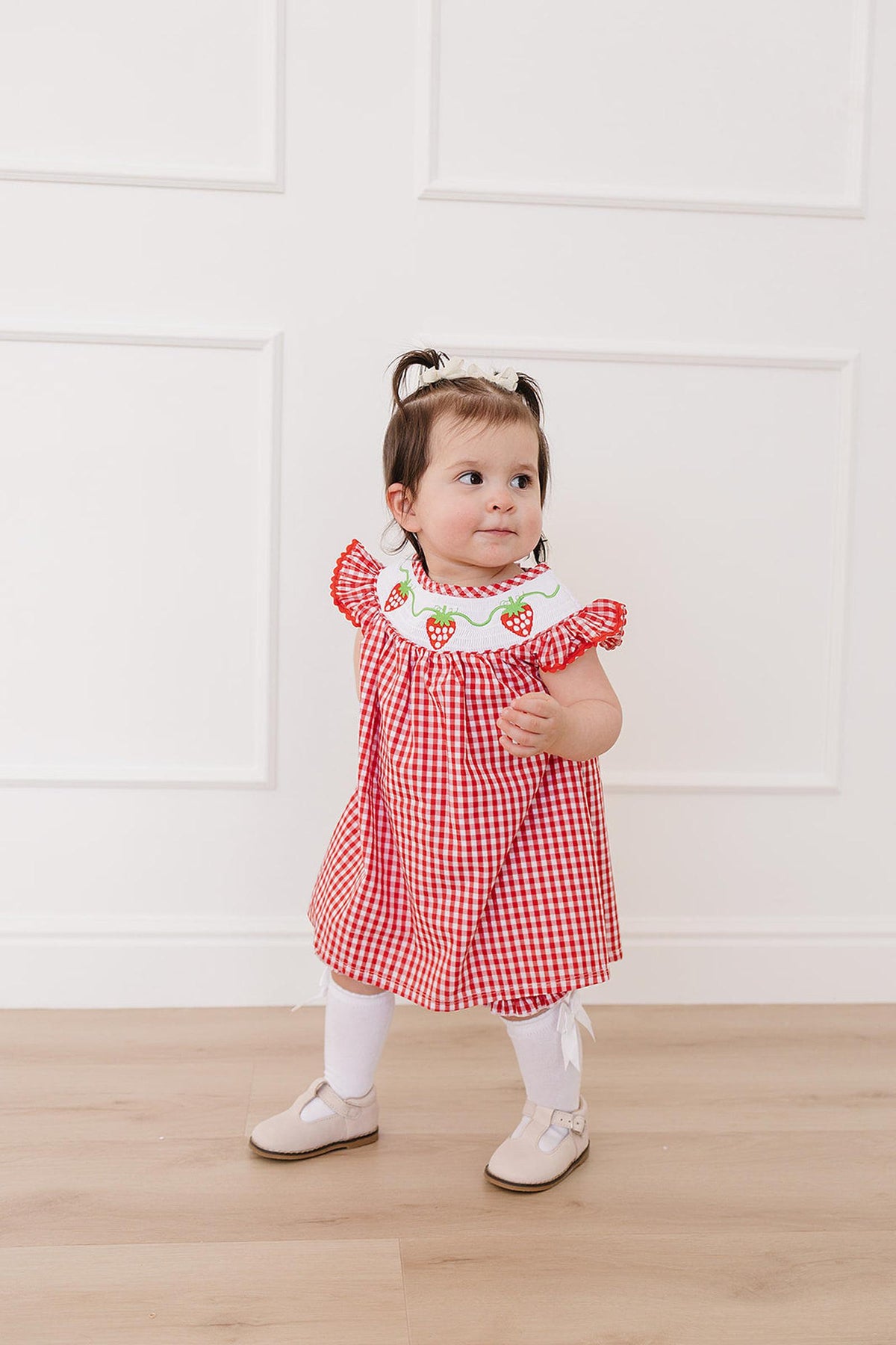 A toddler wears a red and white gingham dress with a strawberry-embroidered collar, paired with white tights and shoes, against a simple, bright background. The outfit is playful and charming.