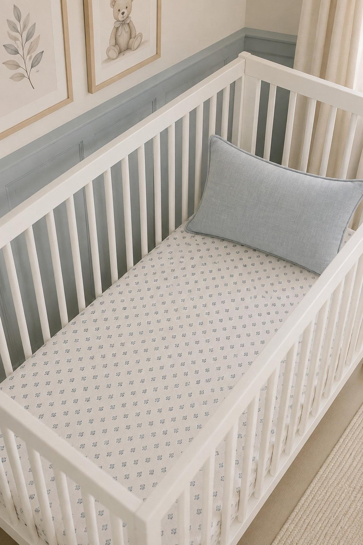 A white wooden crib features a soft muslin crib sheet with a delicate blue floral pattern. The light blue pillow adds a cozy touch to the serene nursery setting.